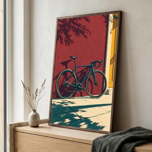 Op de afbeelding: Een ingelijste wandkunstprint met een donkergekleurde racefiets geparkeerd tegen een dieprode muur met een geel deurkozijn. De sc&egrave;ne toont fel zonlicht dat lange, duidelijke schaduwen werpt van de fiets en het nabijgelegen gebladerte.
