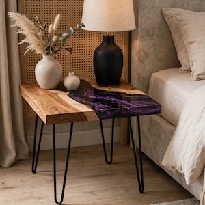 Può includere: Un tavolino quadrato con un piano in legno naturale a bordo vivo e un inserto in resina viola intenso, sostenuto da quattro sottili gambe a forcina in metallo nero. La superficie del tavolo presenta una venatura del legno e una finitura liscia.
