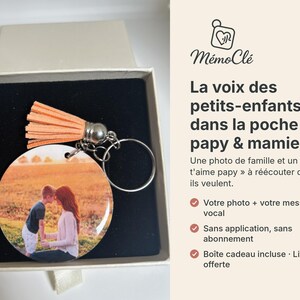 Puede incluir: Un llavero redondo con una fotografía impresa de un niño y un adulto en un campo, unido a una anilla de metal plateado y una borla de color melocotón. El llavero se presenta dentro de una caja cuadrada con un inserto de terciopelo negro.