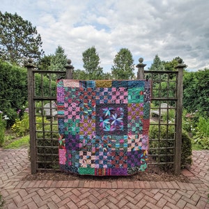 Puede incluir: Una colcha de retazos colorida con una variedad de telas y patrones, incluyendo diseños florales, geométricos y a cuadros. La colcha está colgada en una celosía de madera en un entorno de jardín.