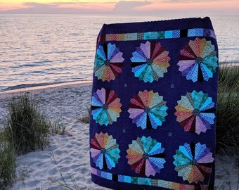 May include: A colorful quilt with a purple background and a pattern of multicolored pinwheels. The quilt is displayed on a sandy beach with a view of the ocean and a sunset in the background.