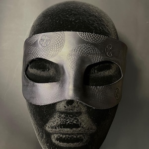 May include: A black leather mask with eye cutouts and embossed floral and scale patterns. The mask is designed to cover the eyes and forehead. The mask is displayed on a black mannequin head.
