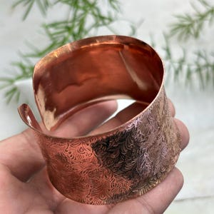 Pode incluir: Uma pulseira larga de cobre com um padrão gravado intrincado de linhas repetitivas em forma de redemoinho e penas na superfície externa. O interior da banda de metal é liso, polido e reflexivo, exibindo um tom metálico quente.