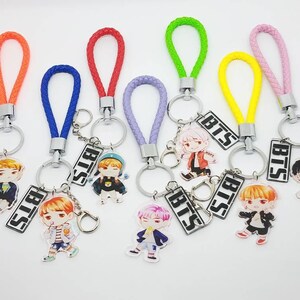 May include: Seven colorful keychains with different colored braided cords and silver key rings. Each keychain features a small, round, printed charm with a cartoon character and the word "BTS" printed on a silver charm.