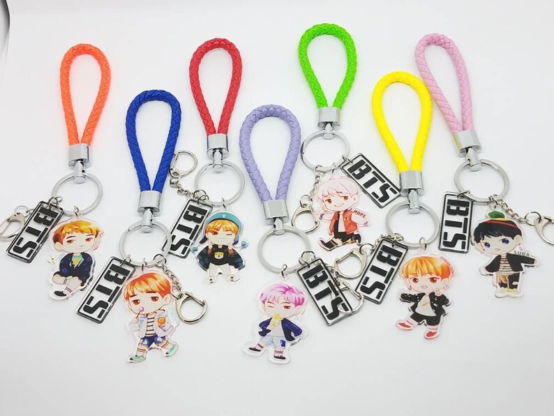May include: Seven colorful keychains with different colored braided cords and silver key rings. Each keychain features a small, round, printed charm with a cartoon character and the word "BTS" printed on a silver charm.