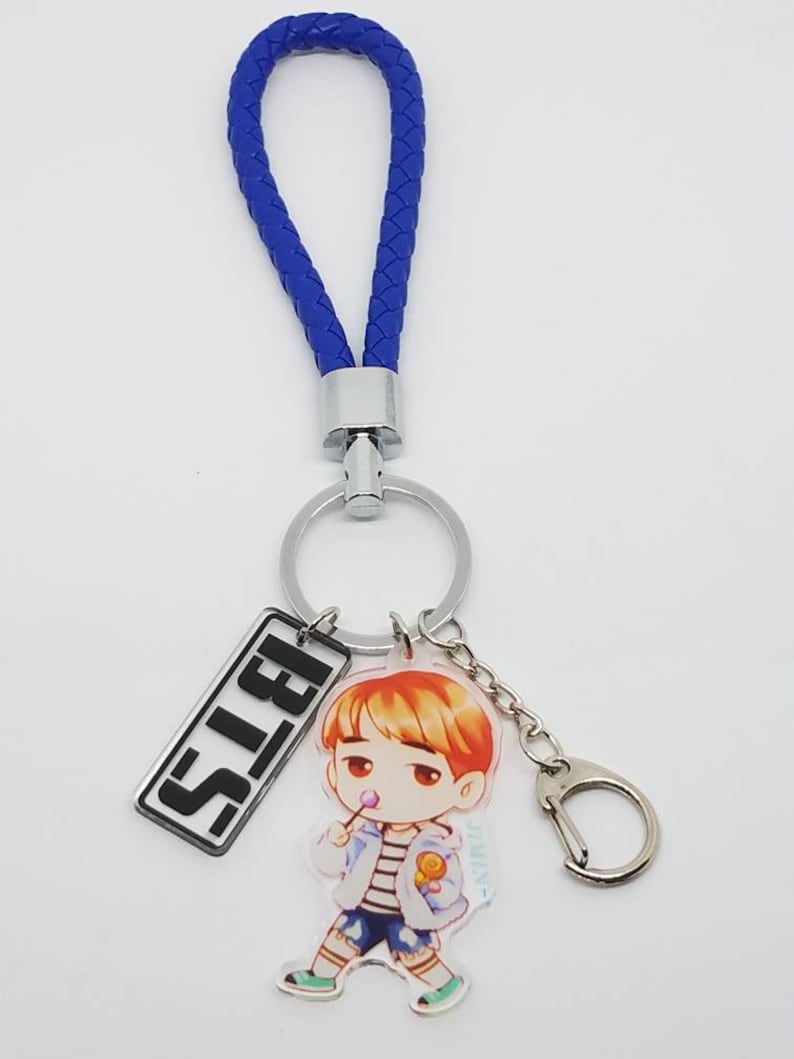 May include: A blue braided keychain with a silver ring and a charm featuring a cartoon character with the word "BTS" on a silver rectangle.