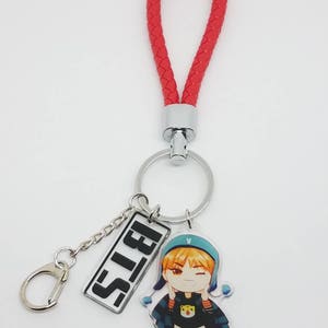 May include: A red braided keychain with a silver ring and a charm featuring a cartoon character with the text "BTS" on a silver rectangle.