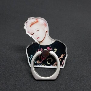 May include: A clear phone ring holder with a silver metal ring. The ring holder features a printed image of a person with pink hair wearing a black shirt with colorful graffiti.