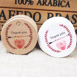 May include: Two round gift tags with a red heart and arrow design. The tag on the left is brown with white text and the tag on the right is white with black text. Both tags say "Thank you - With Love - Oui".