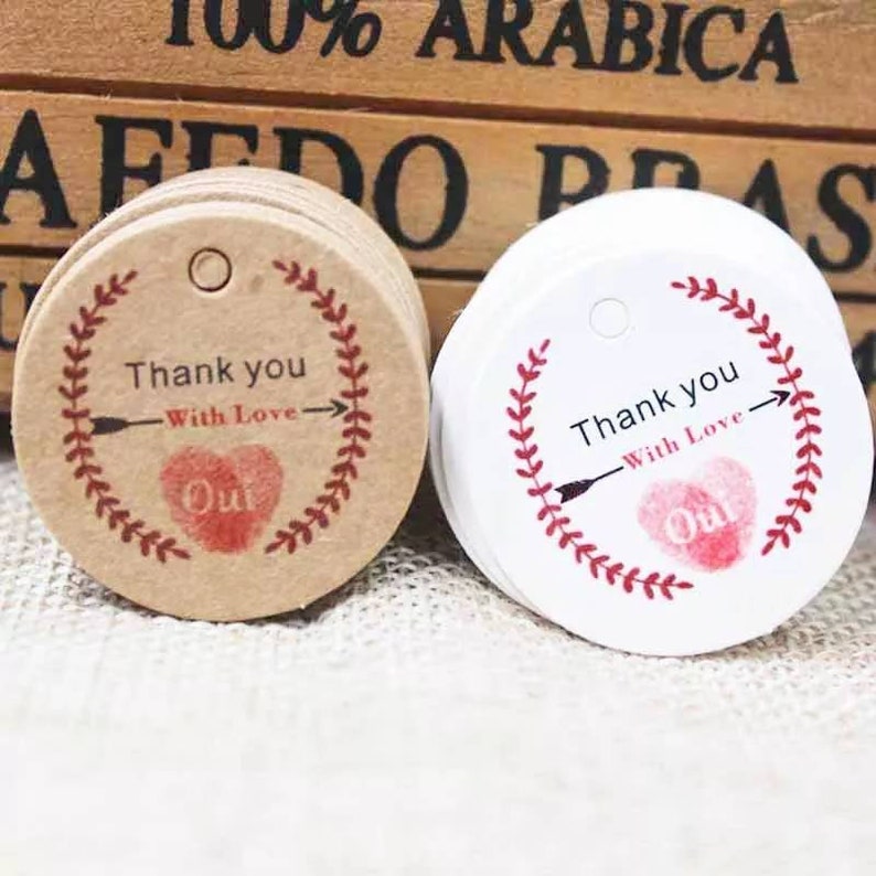 May include: Two round gift tags with a red heart and arrow design. The tag on the left is brown with white text and the tag on the right is white with black text. Both tags say "Thank you - With Love - Oui".