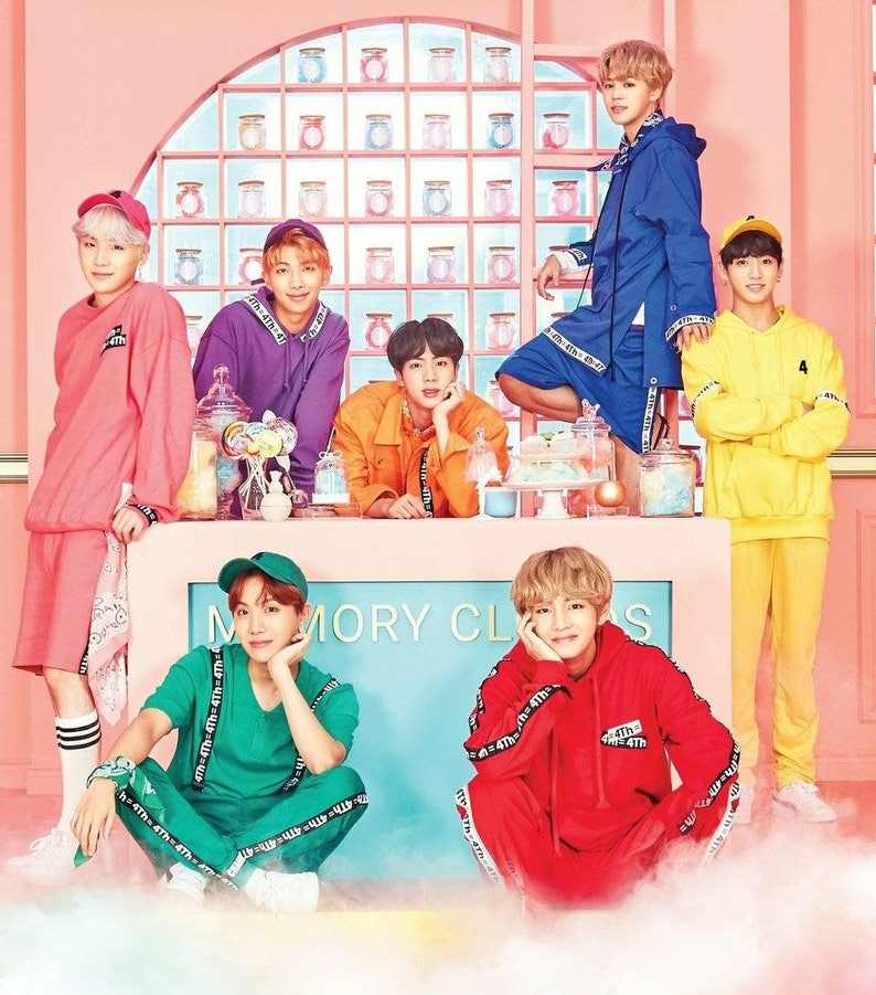 May include: Seven men in colorful clothing pose in front of a pink and white backdrop. The men are wearing various colors, including pink, purple, orange, green, red, blue, and yellow. The backdrop features a wall of jars and the text "MEMORY CLASS".
