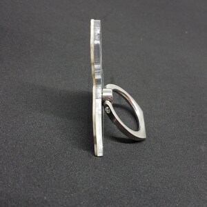 May include: A clear phone ring holder with a silver metal ring. The ring is attached to the holder with a screw.