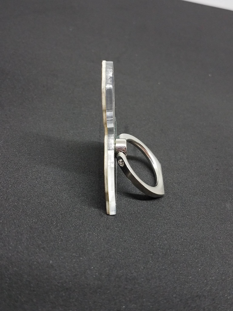 May include: A clear phone ring holder with a silver metal ring. The ring is attached to the holder with a screw.