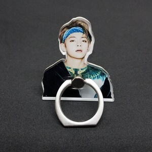 May include: A clear phone ring holder with a silver ring. The holder features a printed image of a person with brown hair wearing a blue bandana and a green and black shirt.
