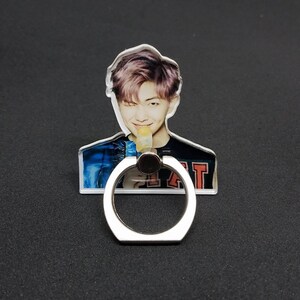 May include: A clear phone ring holder with a photo of a man with purple hair. The man is wearing a blue and black shirt and is holding a yellow popsicle. The ring holder is silver and has a round shape.