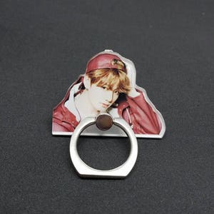 May include: A clear phone ring holder with a silver metal ring. The ring holder features a printed image of a person wearing a red cap and a red jacket.