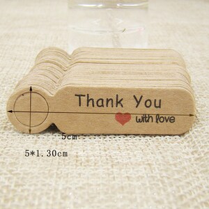 May include: A stack of 50 brown kraft paper gift tags with a hole punched at the top. Each tag has a rounded rectangle shape and is printed with the text "Thank You with love" and a small red heart. The tags measure 5 inches by 1.3 inches.
