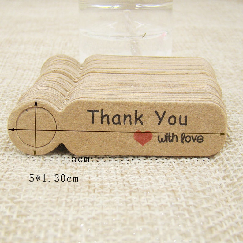 May include: A stack of 50 brown kraft paper gift tags with a hole punched at the top. Each tag has a rounded rectangle shape and is printed with the text "Thank You with love" and a small red heart. The tags measure 5 inches by 1.3 inches.