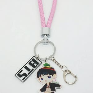 May include: A pink braided keychain with a silver key ring and a charm featuring a cartoon illustration of a person with the text "SUGA" and the word "S.L.E.I."