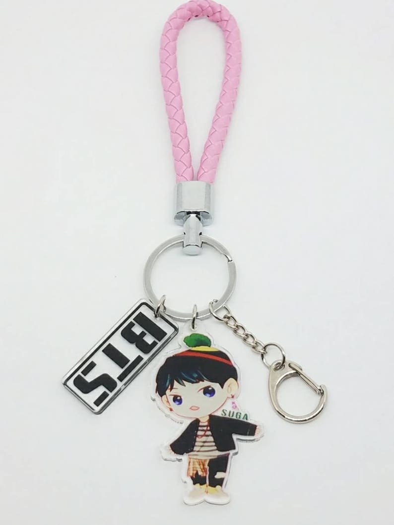 May include: A pink braided keychain with a silver key ring and a charm featuring a cartoon illustration of a person with the text "SUGA" and the word "S.L.E.I."