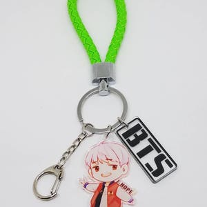 May include: A green braided keychain with a silver ring and a clear acrylic charm featuring a cartoon character with the text "HOPE". A silver charm with the text "BTS" is attached to the ring.