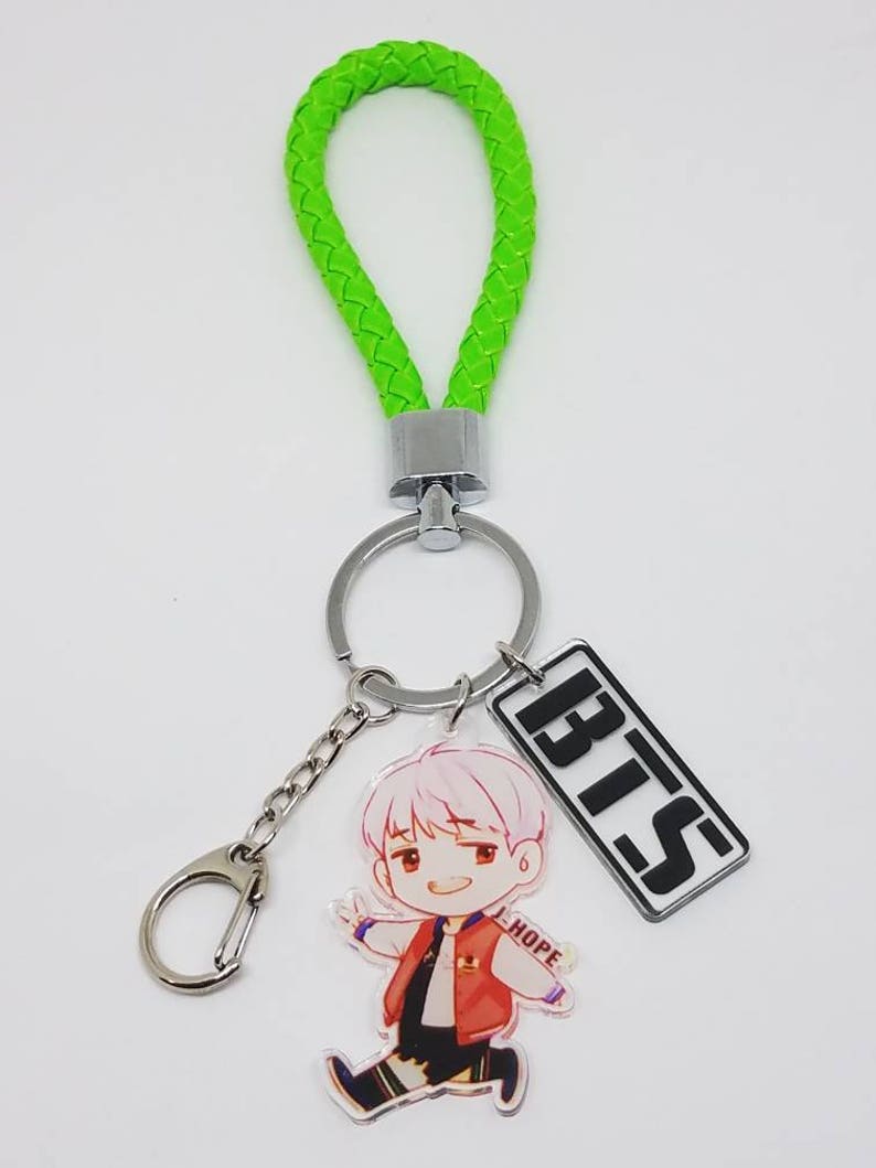 May include: A green braided keychain with a silver ring and a clear acrylic charm featuring a cartoon character with the text "HOPE". A silver charm with the text "BTS" is attached to the ring.