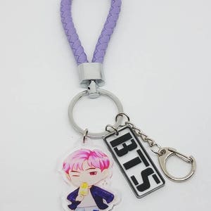 May include: Purple braided keychain with a silver ring and a charm featuring a cartoon character holding an ice cream cone. The charm also includes a silver rectangle with the word "BTS" in black letters.