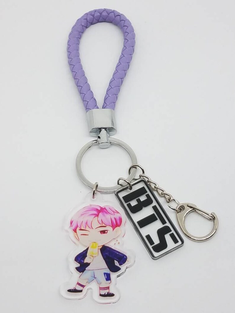May include: Purple braided keychain with a silver ring and a charm featuring a cartoon character holding an ice cream cone. The charm also includes a silver rectangle with the word "BTS" in black letters.