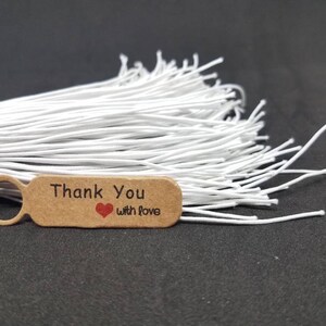 May include: A brown paper tag with a white circle and the text "Thank You with Love" in black lettering. A red heart is printed between the words "Thank" and "with". The tag is attached to a bundle of white string.