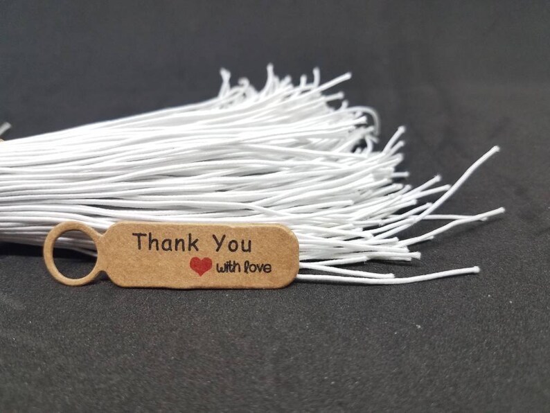 May include: A brown paper tag with a white circle and the text "Thank You with Love" in black lettering. A red heart is printed between the words "Thank" and "with". The tag is attached to a bundle of white string.
