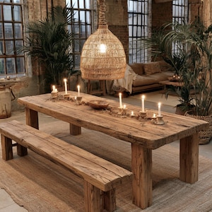 May include: A long, rectangular, weathered wooden dining table with a matching bench. The table is decorated with multiple lit white taper candles in holders and a small wooden bowl. A large, woven, bell-shaped pendant light hangs above.