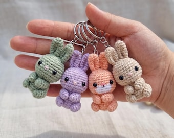 May include: Four small crochet rabbit keychains in sage green, lavender, peach, and beige. Each stuffed animal features black bead eyes, a small embroidered nose, and a metal split ring attached to the top of its head for hanging.