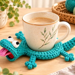 Puede incluir: Un posavasos de ganchillo turquesa con forma de lagarto y una taza de cerámica blanca con motas marrones sobre su espalda. El lagarto tiene una lengua rosa, ojos blancos con pupilas negras y cuatro patas pequeñas. Se muestran etapas del tejido.