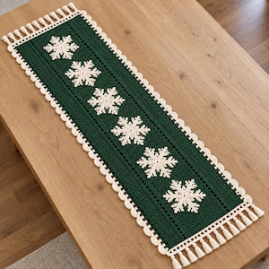 May include: A rectangular dark green crochet table runner featuring six cream-colored snowflake motifs arranged in a row. The runner is bordered by a scalloped cream edge and finished with small cream tassels at each of the four corners.