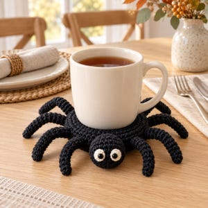 May include: A black crocheted spider coaster with eight legs and two white eyes with black pupils, supporting a plain white ceramic mug filled with a brown beverage. The spider is crafted from yarn with a circular body and textured surface.