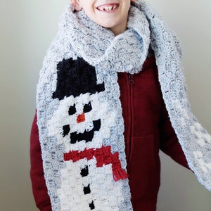 Puede incluir: Un niño con una sudadera roja y una bufanda de ganchillo gris con un diseño de muñeco de nieve. El muñeco de nieve es blanco con ojos negros, boca negra, nariz roja y bufanda roja.