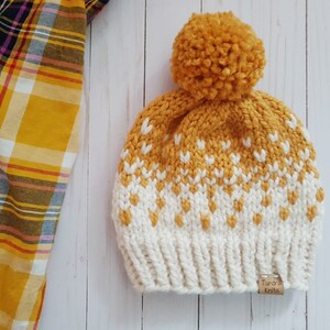 May include: A yellow and white knitted hat with a pom pom on top. The hat has a fair isle pattern and a brown label that says "Tundra Knits".