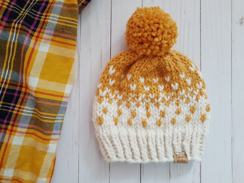May include: A yellow and white knitted hat with a pom pom on top. The hat has a fair isle pattern and a brown label that says "Tundra Knits".