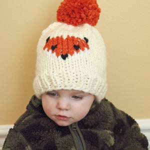 May include: A white knitted hat with a fox face design in orange and black. The hat has a fluffy orange pom-pom on top.