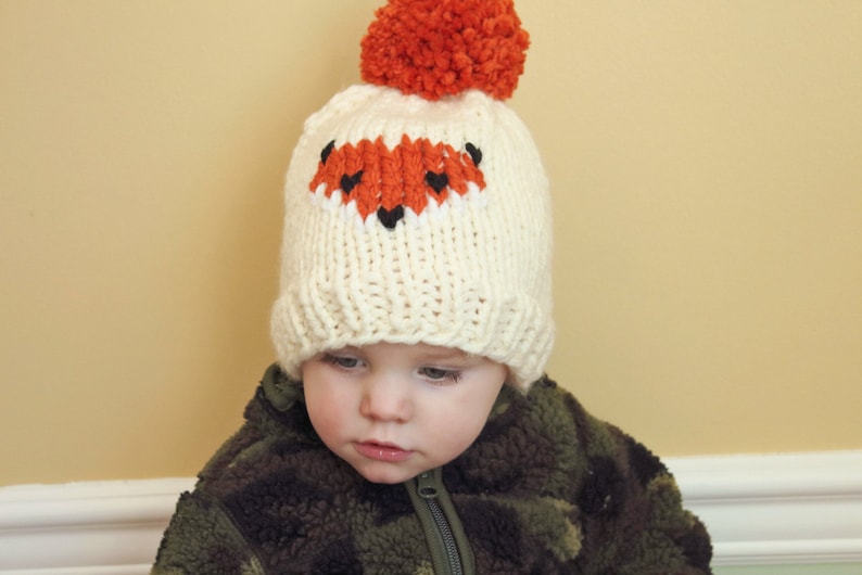 May include: A white knitted hat with a fox face design in orange and black. The hat has a fluffy orange pom-pom on top.