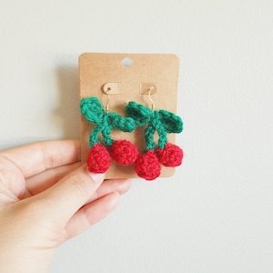 Puede incluir: Un par de pendientes de cereza de ganchillo con hojas verdes y cerezas rojas. Los pendientes están en una tarjeta marrón.