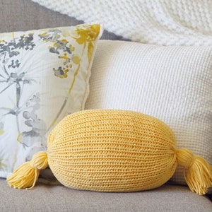 May include: A yellow knitted oval pillow with two tassels on either end. The pillow is sitting on a gray couch with other pillows behind it.