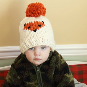 May include: A toddler wearing a white knitted hat with an orange fox face design and a brown and green camouflage jacket.