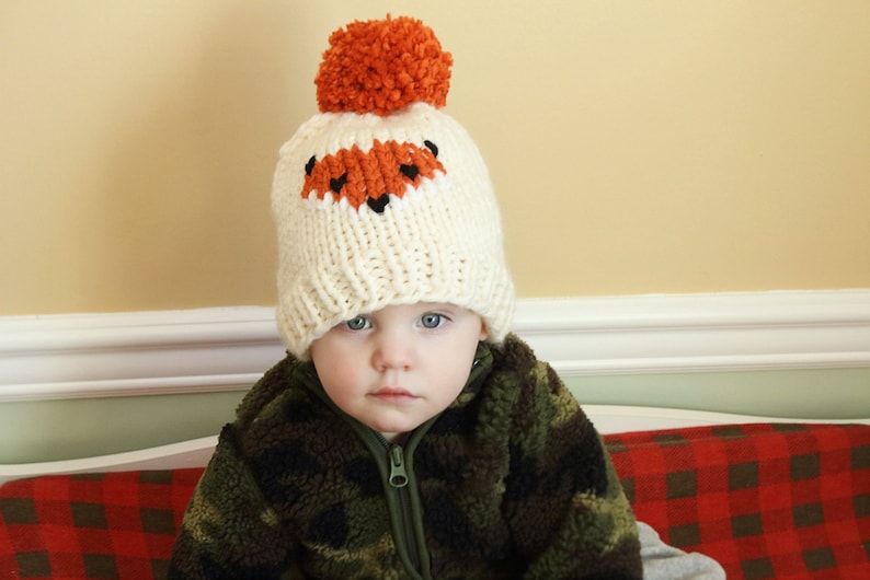 May include: A toddler wearing a white knitted hat with an orange fox face design and a brown and green camouflage jacket.