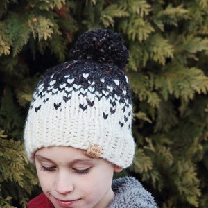 May include: A white and black knit beanie with a black pom pom on top. The beanie has a heart pattern and a brown leather tag that says " Tundra Knits".