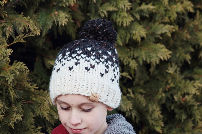 May include: A white and black knit beanie with a black pom pom on top. The beanie has a heart pattern and a brown leather tag that says " Tundra Knits".