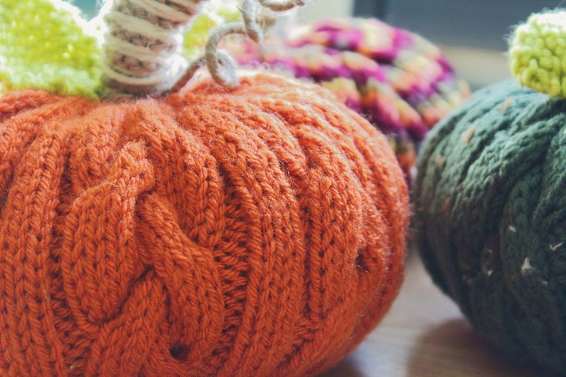 Puede incluir: Tres calabazas de punto en naranja, verde y morado. Las calabazas est&aacute;n hechas con punto de cable y tienen un aspecto texturizado.