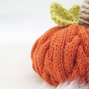 Puede incluir: Una calabaza naranja de punto con una hoja verde de ganchillo y un tallo marr&oacute;n. La calabaza est&aacute; sentada sobre una superficie blanca y esponjosa.