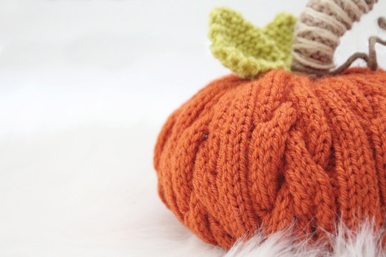 Puede incluir: Una calabaza naranja de punto con una hoja verde de ganchillo y un tallo marr&oacute;n. La calabaza est&aacute; sentada sobre una superficie blanca y esponjosa.