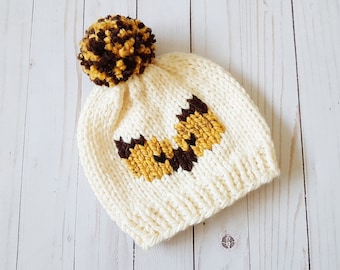 May include: A white knitted hat with a brown and yellow bee design. The hat has a brown pom-pom on top.